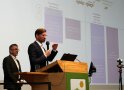 Mann steht an Rednerpult, dahinter weiterer Mann