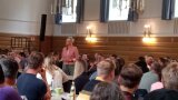 Person steht in einem Saal zwischen Personen an Tischen 