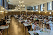 Personen sitzen an Einzeltischen in einem Saal und schreiben